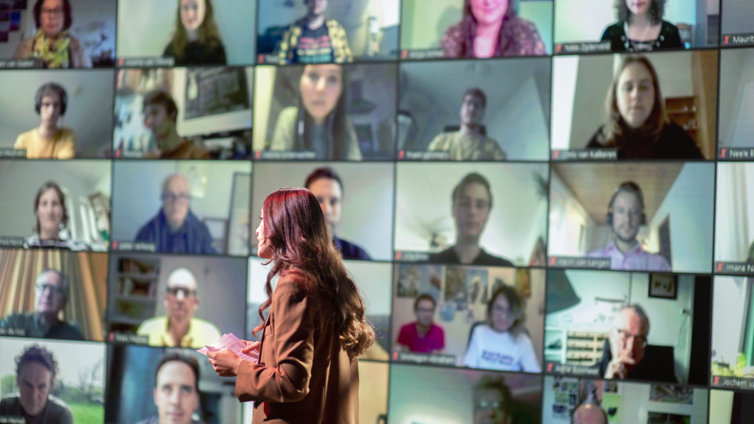 Emmelie, een vrouw met lang donker haar, staat voor een muur met een videoconferentie met allerlei mensen erop.